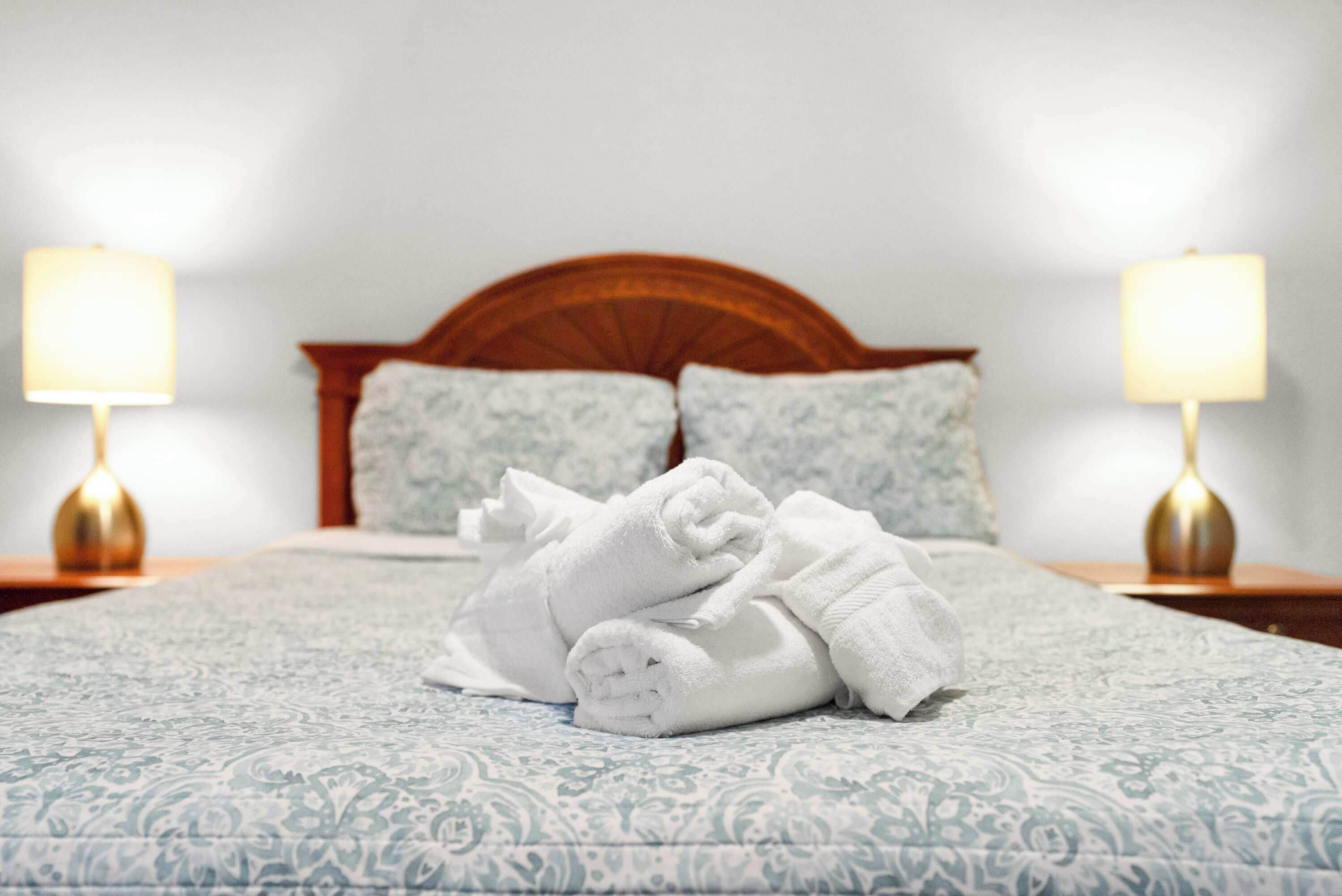 Een net opgemaakt bed met een beddekleed met dessin, twee kussens en een stapel opgevouwen handdoeken op het bed.