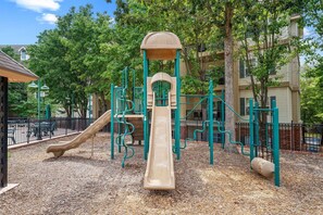 Ein Spielplatz mit Rutsche und Klettergerüsten, umgeben von Bäumen und einem Zaun.