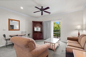Quarto de hotel com ventoinha no teto, mesa de jantar com cadeiras, sofá, pequena mesa com abajur, espelho e vista para o exterior pela janela.