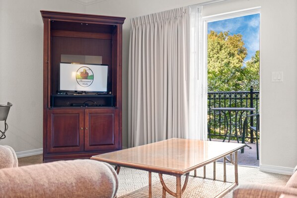 Quarto com televisão sobre um armário de madeira, uma mesa e cadeiras. Há uma varanda com vista para árvores e um corrimão.