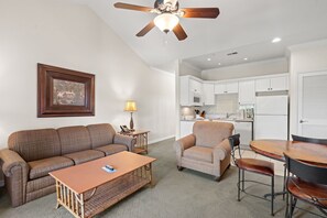A living room with a sofa, coffee table, and armchair, a dining table with chairs, a ceiling fan, and a kitchen area with white cabinets and appliances. - Resort King Condo | Balcony • Pool • Hot Tub (Branson)