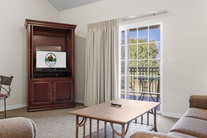 A room with a television, a wooden cabinet, a table, and a view of a balcony with a railing. - Resort King Condo | Balcony • Pool • Hot Tub (Branson)