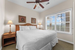 A bedroom with a bed, two nightstands, two wall-mounted lamps, a ceiling fan, and a large window with blinds. - King Condo Near Silver Dollar City & Highway 76 (Branson)