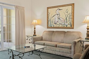 A living room with a beige sofa, a glass coffee table, two lamps, and a framed painting of birds on the wall. - Family Condo | 2 Kings, Loft | Great Location (Branson)