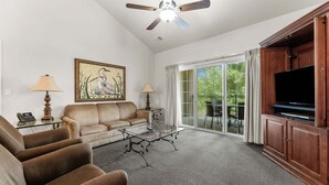 A living room with a sofa, armchairs, a coffee table, a painting, a ceiling fan, a television, and a sliding glass door leading to a patio. - Spacious Family Condo | 2 Kings + Loft (Branson)