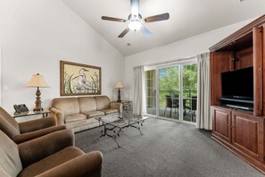 Ein Wohnzimmer mit Sofa, Sesseln, Couchtisch, einem Gemälde, Deckenventilator, Fernseher und einer Schiebetür zu einer Terrasse.