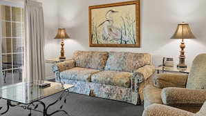 Sala de estar com sofá, duas poltronas, mesa de centro de vidro e duas luminárias. Há um quadro de um pássaro na parede.