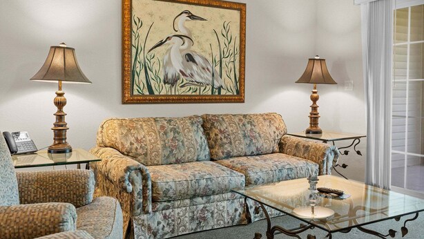 A living room with a sofa, two armchairs, a glass coffee table, and two lamps. A framed painting of a bird hangs on the wall. - French Quarter Resort | Family Condo Near Shows (Branson)