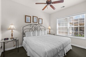 Ein Schlafzimmer mit einem Bett, zwei Nachttischen, einem Deckenventilator, einem Fenster mit Jalousien und gerahmten Bildern an der Wand.
