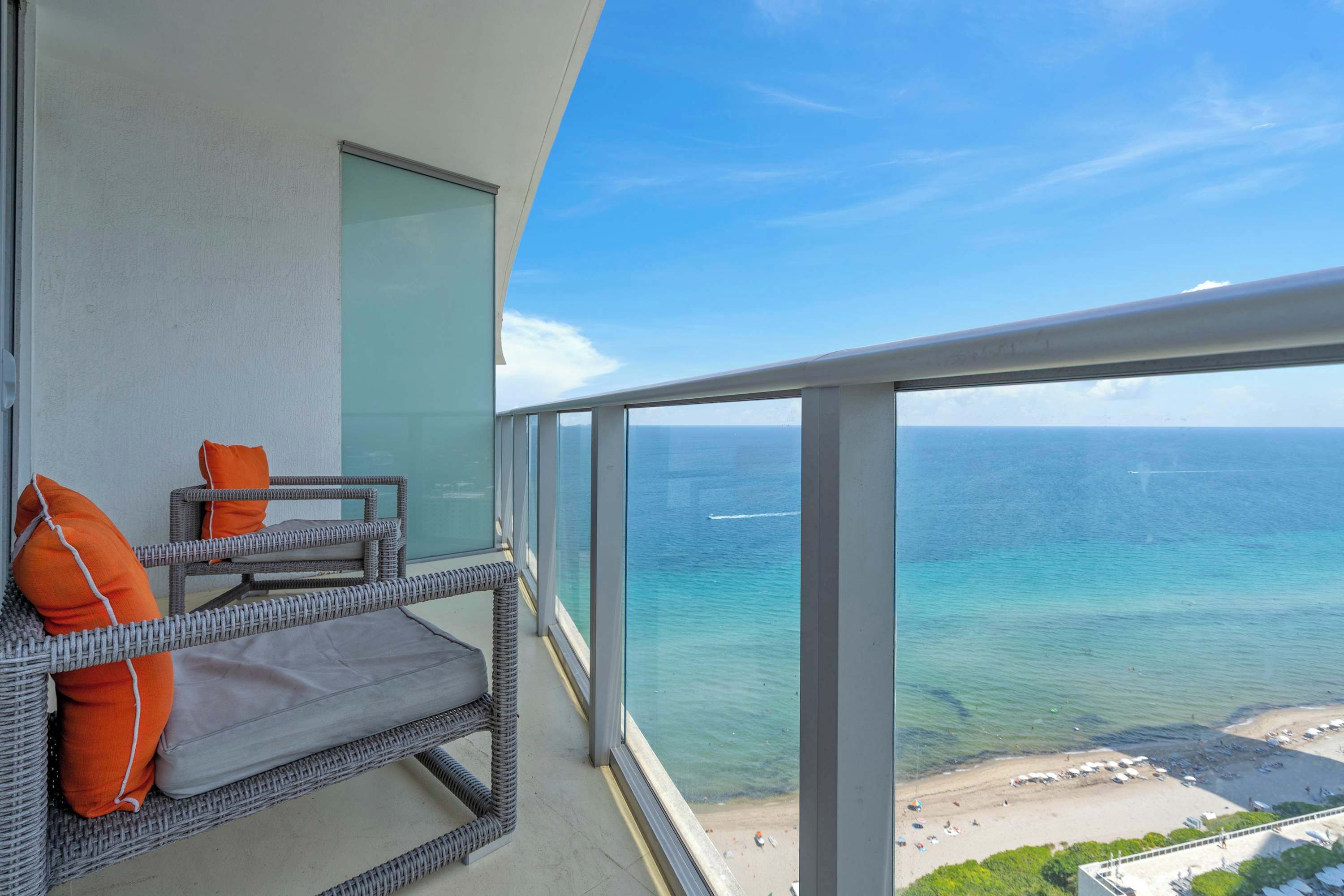 A balcony with wicker furniture and orange cushions, overlooking a beach and the ocean.