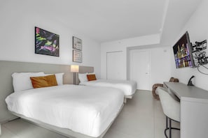 Ein Hotelzimmer mit zwei Betten, einem Schreibtisch und einem Stuhl. An den Wänden befinden sich Kunstwerke, und eine Tür führt in einen weiteren Raum.