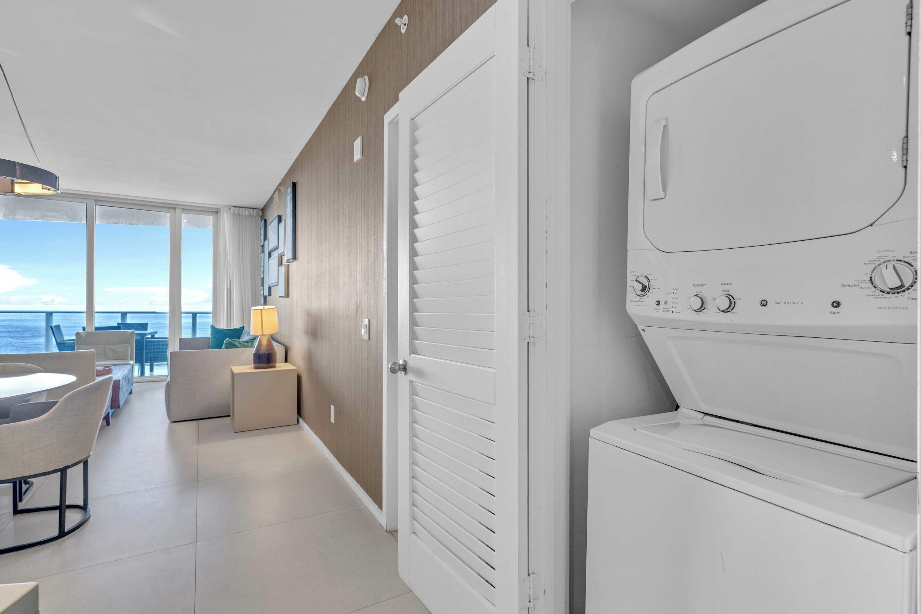 A hotel room with a large window, a washing machine, and a dining area with a table and chairs.