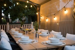 An outdoor dining area with tables set for a meal, string lights overhead, and a view of trees in the background.