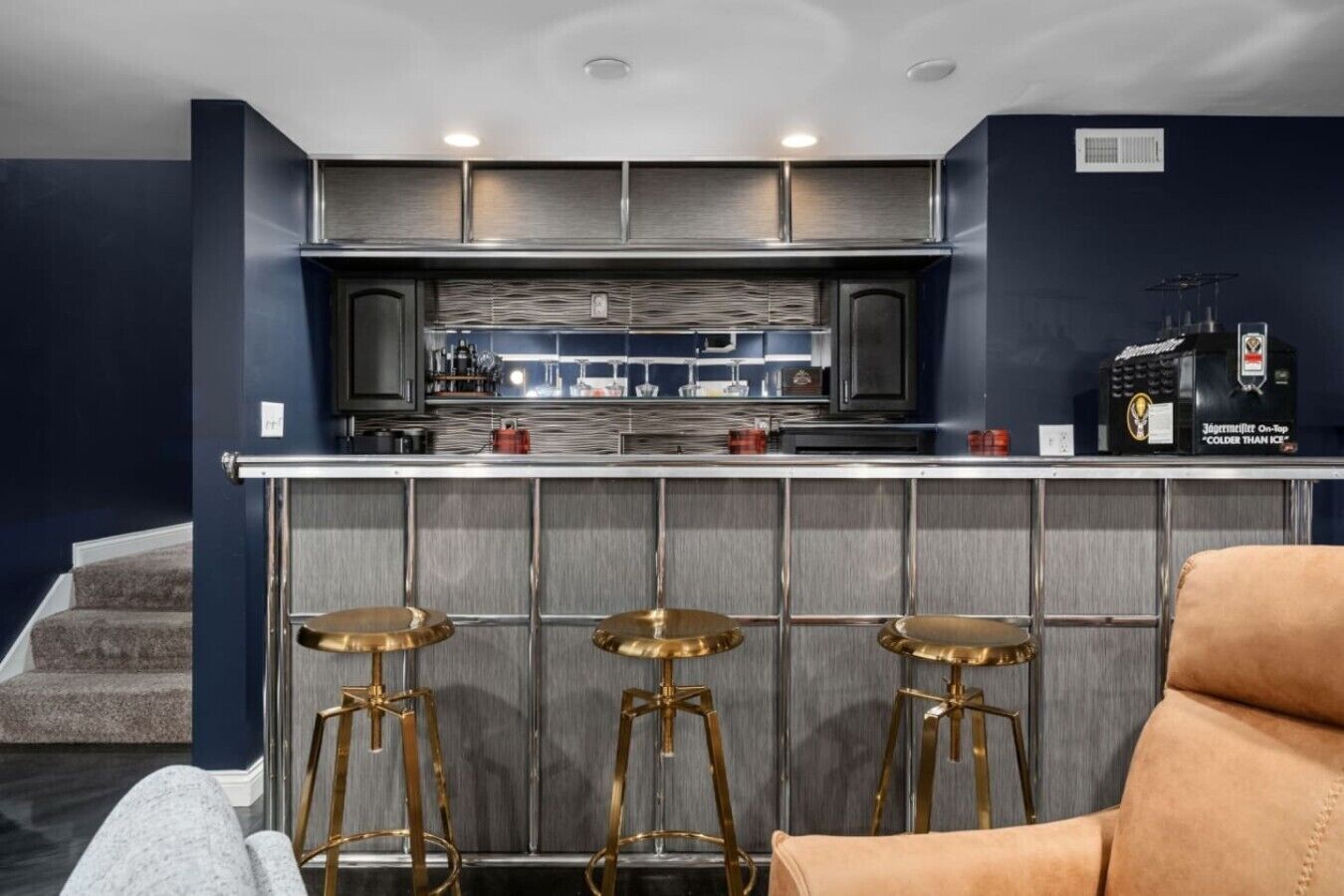 A modern kitchen with a bar area, featuring stainless steel appliances, a bar stool setup, and a visible refrigerator.