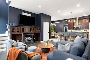 A modern living room with a fireplace, seating area, and a kitchen in the background. - Soulard Sensational|Sleeps18|HotTub|PuttPutt|Games (St. Louis)