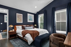 A bedroom with a large bed, bedside lamps, a wooden nightstand, a bench, and a window with curtains. - Soulard Sensational|Sleeps18|HotTub|PuttPutt|Games (St. Louis)