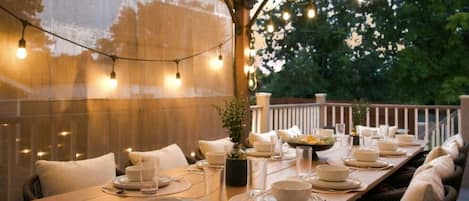 An outdoor dining area with a long wooden table set for a meal, string lights overhead, and a view of trees in the background.