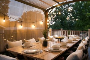 An outdoor dining area with a long wooden table set for a meal, string lights overhead, and a view of trees in the background.