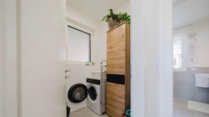 Ein kleiner Waschraum mit Waschmaschine und Trockner, einem Holzkasten mit einer Pflanze und Blick in ein Badezimmer mit Handtuchhalter.