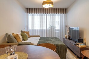 Ein Hotelzimmer mit Bett, Fernseher, Esstisch und einem großen Fenster mit Vorhängen.