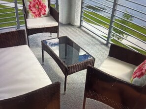 Ein Balkon mit Korbmöbeln, einem Glastisch und Blick auf eine Rasenfläche.