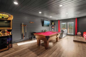 A pool table in a room with wooden flooring, a television mounted on the wall, and a pool table with a red felt top. - Luxe Themed Cabin | Theater | Hot Tub | Mtn Views | Sleeps 18+ (Pigeon Forge)