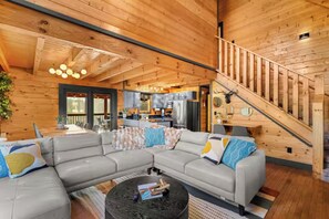 A spacious living room with a large sectional sofa, a coffee table, and a staircase leading to an upper level. - Luxe Themed Cabin | Theater | Hot Tub | Mtn Views | Sleeps 18+ (Pigeon Forge)