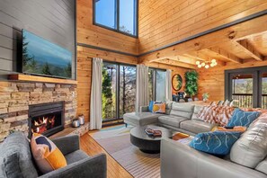 Una sala moderna con chimenea de piedra, un sofá modular y un amplio ventanal con vistas a un bosque.