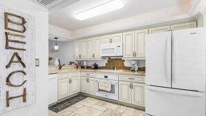 Una cocina con armarios blancos, un microondas, un refrigerador y un adorno de pared de madera con la palabra 'BEACH'.