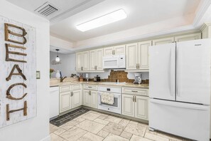 A kitchen with white cabinets, a microwave, a refrigerator, and a wooden wall decoration spelling 'BEACH'. - Coastal Condo with Pool, Spa & Beach Fun Awaits - Condo Islands End 602 - Roelens (Fort Myers Beach)