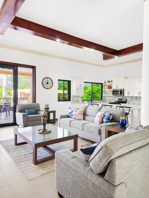 A modern living room with a sofa, coffee table, and a dining area with chairs. There is a kitchen with white cabinets and a microwave. - Spacious 12 Bedrooms Villa for Large Groups in St Ann, Jamaica (Saint Ann's Bay)
