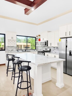A modern kitchen with white cabinetry, a marble countertop, a stainless steel refrigerator, and a large window offering a view of greenery. - Spacious 12 Bedrooms Villa for Large Groups in St Ann, Jamaica (Saint Ann's Bay)