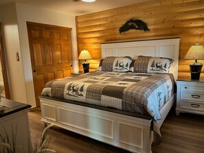 Une chambre à coucher avec une tête de lit en bois, une courtepointe ornée de motifs de cerfs et de forêt, deux lampes de chevet et une porte en bois.