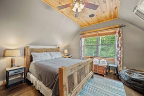 Ein Schlafzimmer mit einem Holzbett, einem Nachttisch, einer Lampe, einem Fenster mit Vorhängen und einem Deckenventilator.