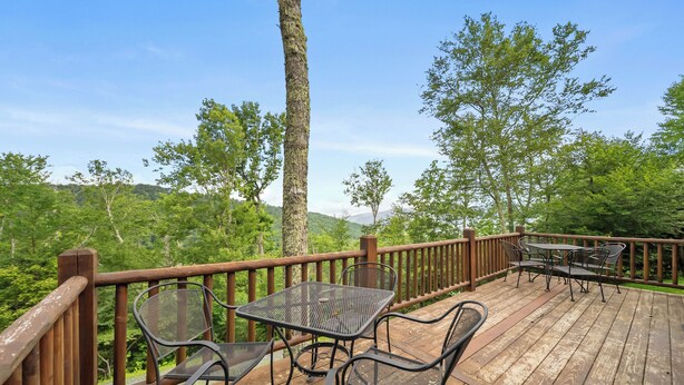 Eine Holzterrasse mit Metallstühlen und einem Tisch, umgeben von einer Holzbrüstung, mit Blick auf eine bewaldete Landschaft mit Bäumen und einem Berg in der Ferne.
