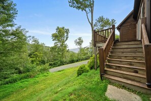 A wooden cabin with a staircase leading to a deck, surrounded by lush greenery and a gravel path. - Lazy Daze: Views, Pet Friendly, Outdoor Pavilion (Banner Elk)