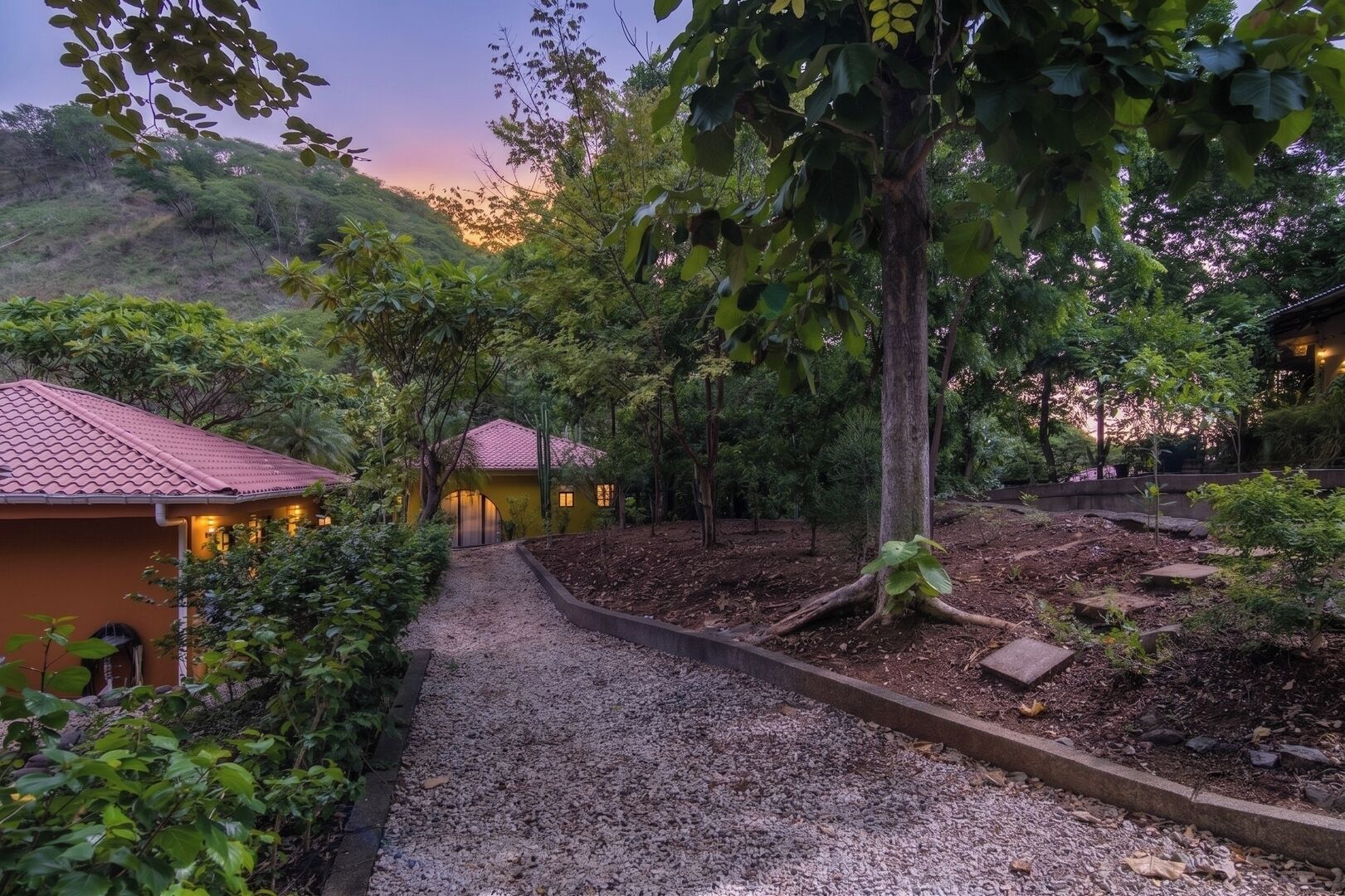 Un chemin menant à un bâtiment dont le toit est recouvert de tuiles rouges, entouré d’une végétation luxuriante et d’arbres.