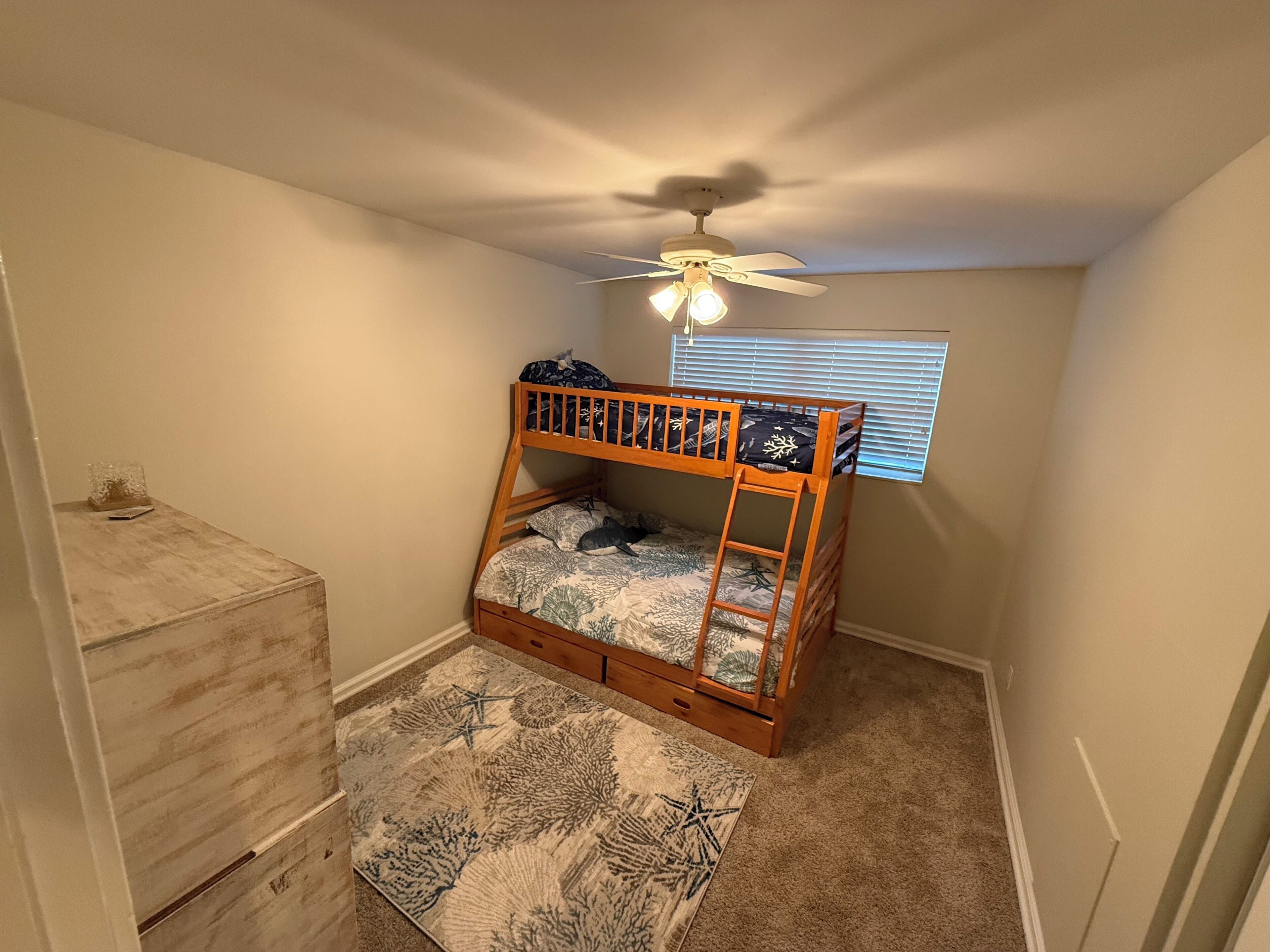 A small, single-bed room with a wooden bunk bed, a ceiling fan, a window with blinds, a wooden dresser, and a patterned rug.