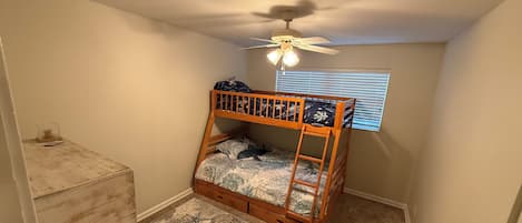 A small, single-bed room with a wooden bunk bed, a ceiling fan, a window with blinds, a wooden dresser, and a patterned rug.