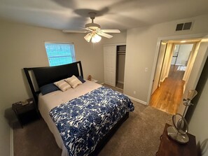 Ein Schlafzimmer mit einem Bett, einem Deckenventilator, einem Fenster mit Jalousien, einem Kleiderschrank und einem Flur, der zu einem weiteren Zimmer führt.