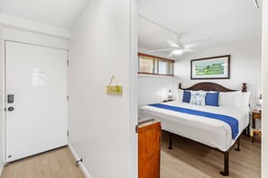 Ein Schlafzimmer mit einem Bett, einem Deckenventilator, einem Fenster mit Jalousien, einem Nachttisch mit Lampe und einem gerahmten Bild an der Wand.