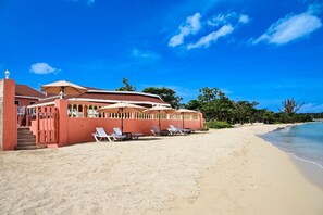 Un edificio frente a la playa con fachada roja, mobiliario exterior con sombrillas y una playa de arena.