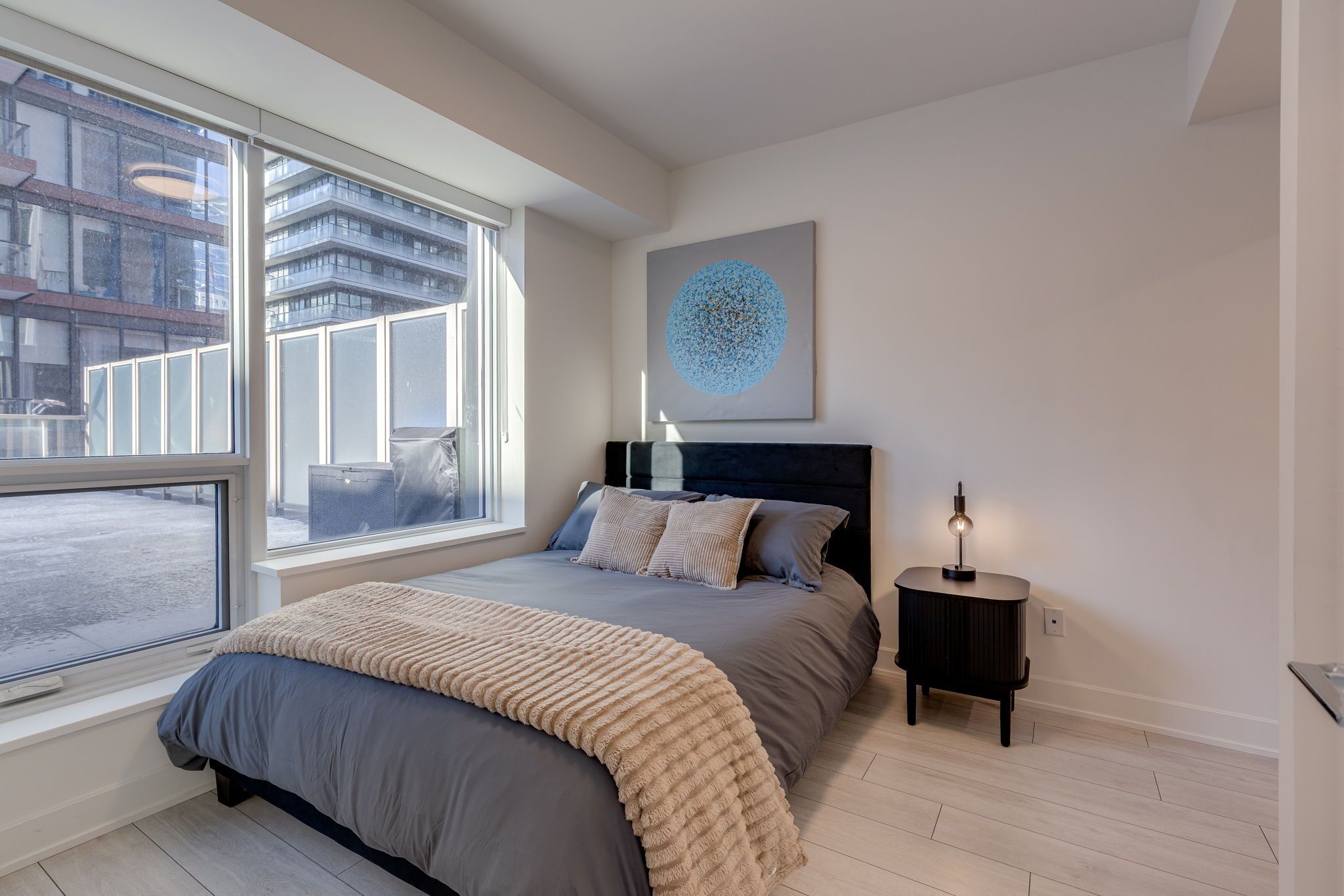 A modern bedroom with a large window, a bed with a grey and beige duvet, a black headboard, a bedside table with a lamp, and a wall art piece.