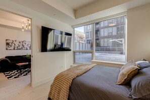 Ein modernes Schlafzimmer mit einem großen Bett, einem flachbildfernseher an der Wand und Blick auf die Stadt durch große Fenster.