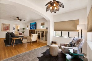 A living room with a sofa, armchair, ottoman, and a coffee table. There is a ceiling fan, a wall-mounted TV, and a window with blinds. - Old Town Buffalo+Private Hot Tub+Near Old Town+Pet Friendly! (Albuquerque)