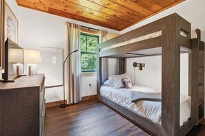 Una habitació amb un llit superposat, un escriptori amb una làmpada, una finestra amb cortines i vista a vegetació a l'exterior.