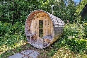 Eine zylindrische Holzhütte mit Tür und Sitzbank im Inneren, umgeben von viel Grün.