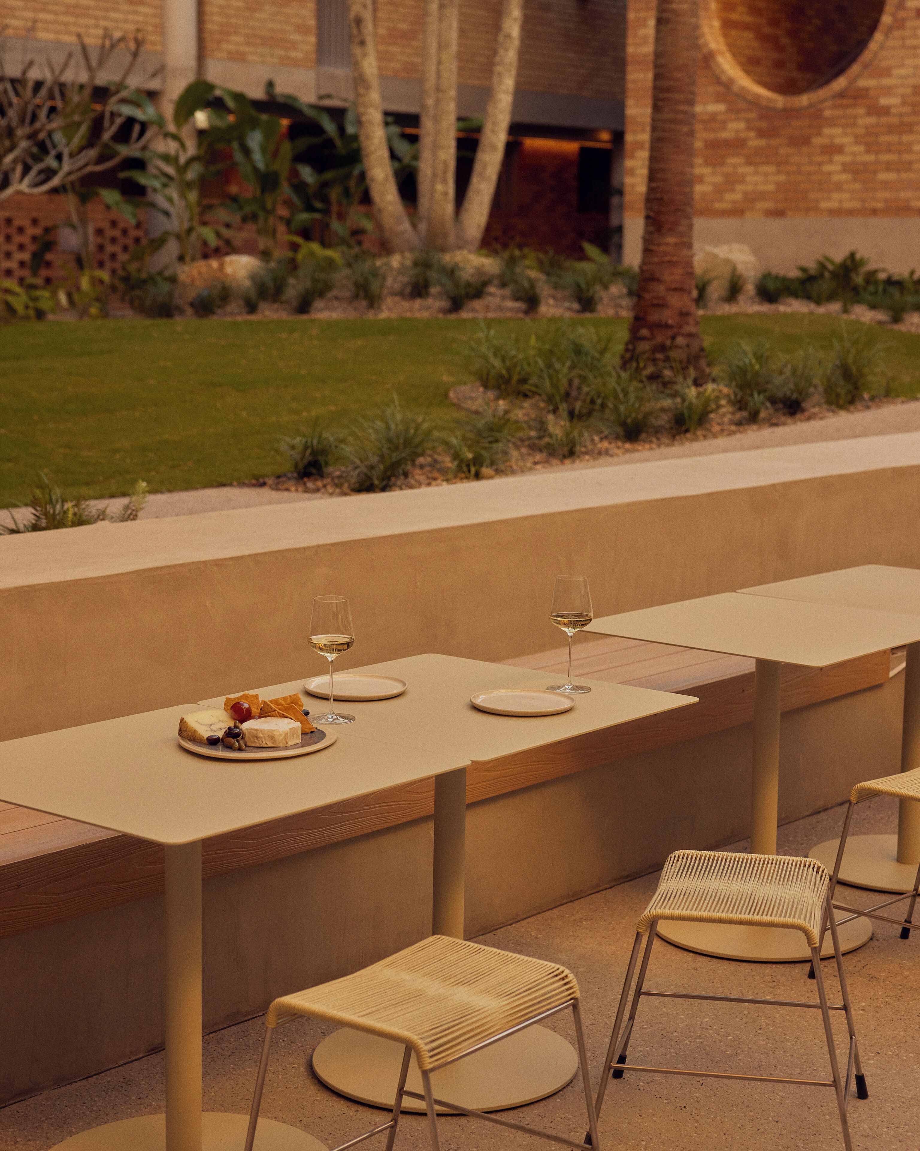 Un espacio de comedor al aire libre con una mesa larga y cuatro sillas, rodeado por un jardín con plantas y un muro de ladrillo.