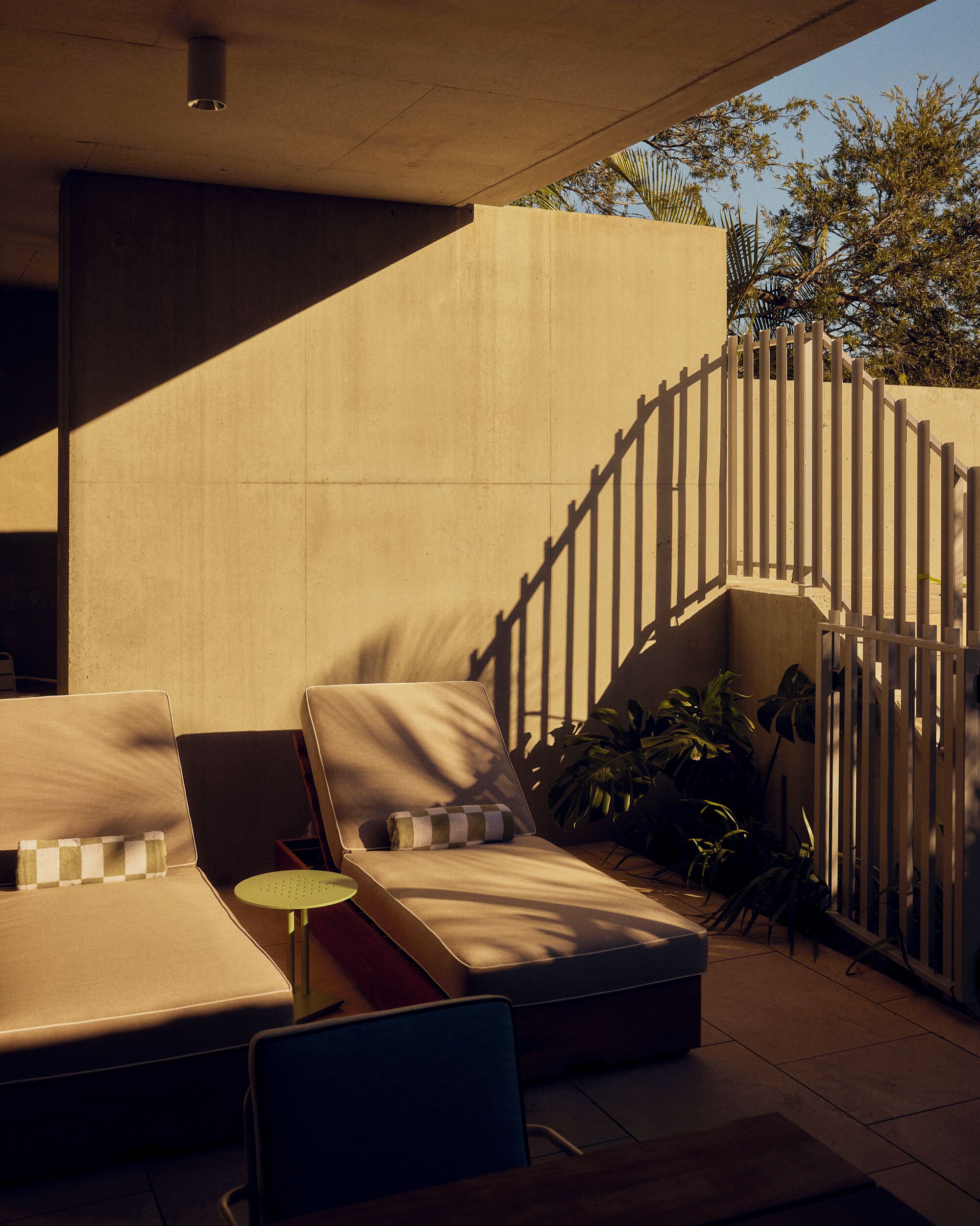 A sunlit outdoor seating area with lounge chairs and a small table, a concrete wall, and a wooden fence.
