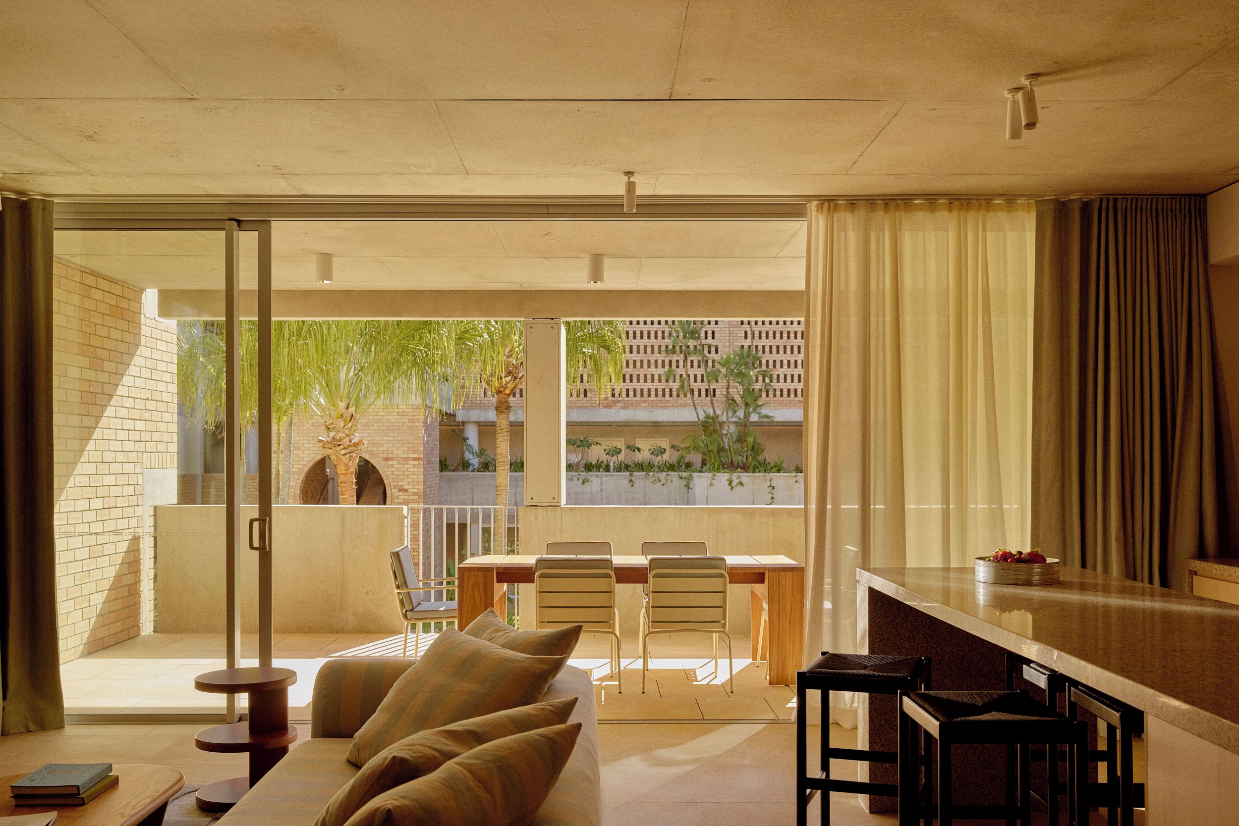 A modern living room with a sofa, a dining table with chairs, and a view through a glass door to an outdoor area with plants and a brick wall.